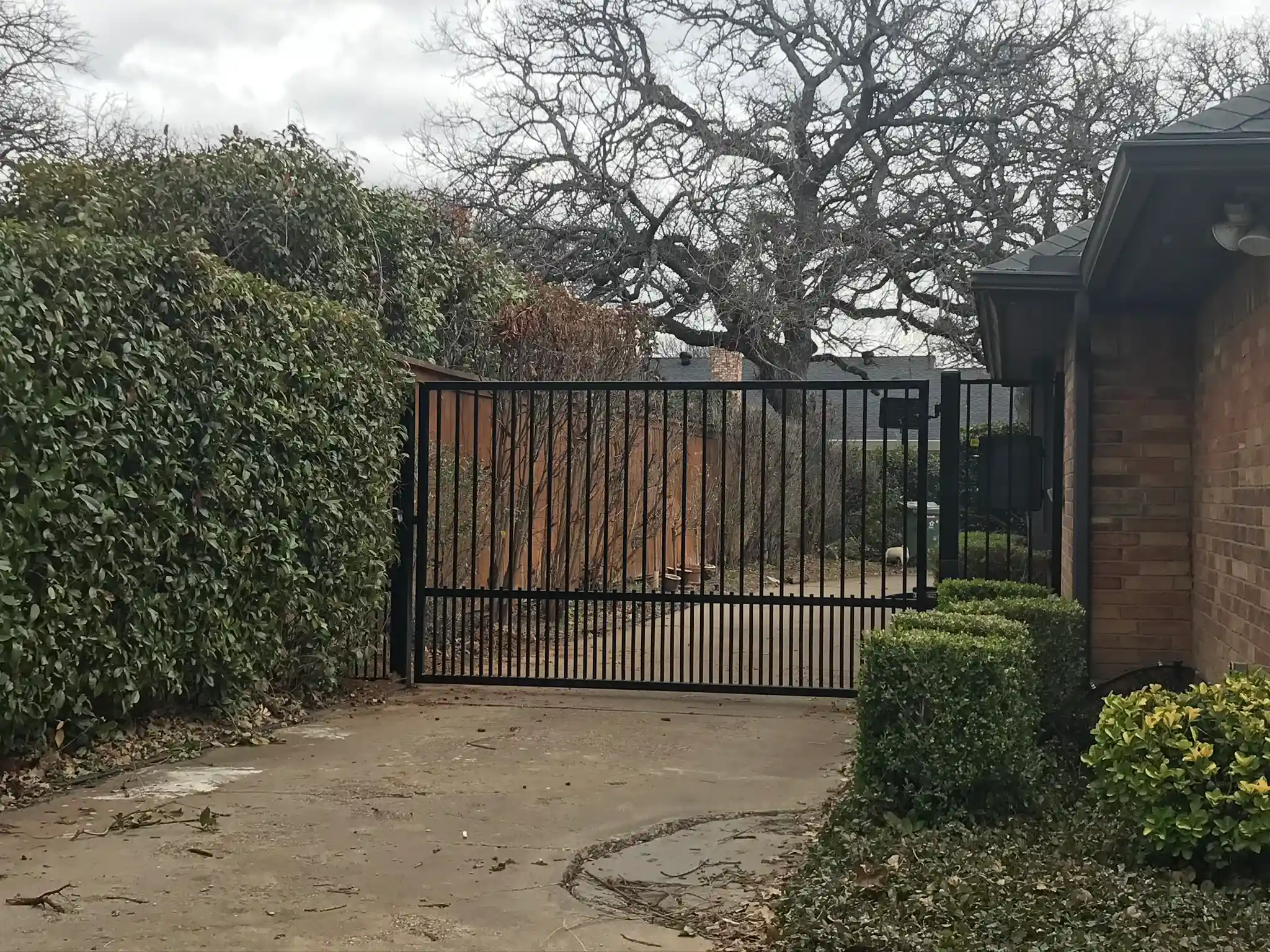 Flat-top black steel driveway gate with vertical pickets, installed between a tall green hedge and a brick home